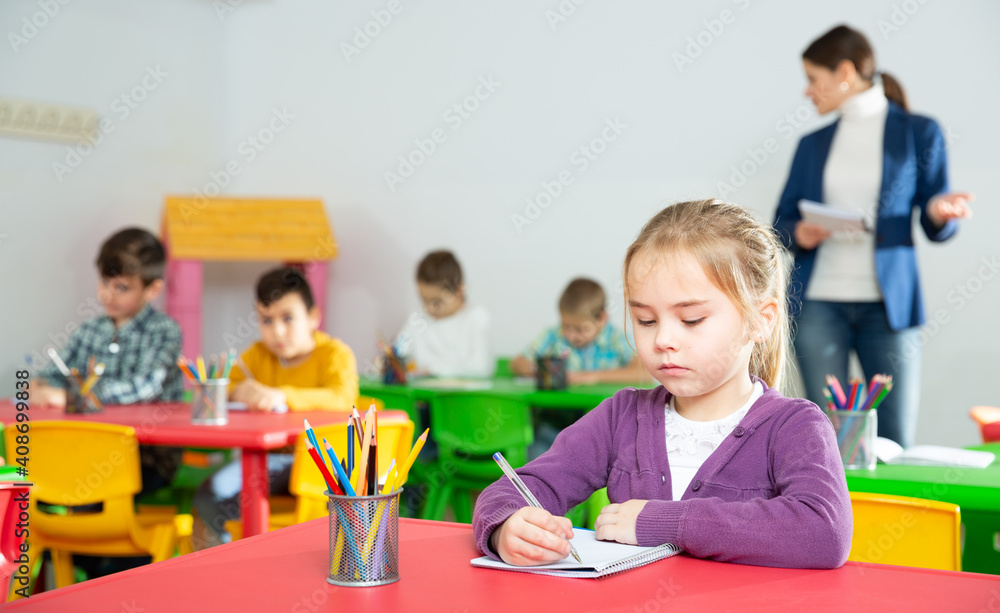 Portrait of assiduous little girl with pen and notebook at lesson in elementary school - obrazy, fototapety, plakaty 