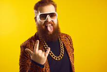 Studio Portrait Of Funny Eccentric Rich Bearded Chubby Young Man In Sunglasses, Extravagant Funky Outfit And Huge Gold Chain Around Neck Doing Rock Horn Sign, Looking At Camera And Sticking Tongue Out