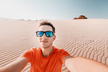 Happy Man Tramp In Sunglasses Takes A Selfie Photo In The Desert Against The Background Of A Dune Of White Sand