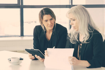 Wall Mural - Serious businesswomen discussing reports. Two female professionals sitting together, holding documents, using tablet and talking. Communication concept