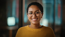 Close Up Portrait Of A Young Latina With Short Dark Hair And Glasses Posing For Camera In Creative Office. Beautiful Diverse Multiethnic Hispanic Female Wearing Yellow Jumper Is Happy And Smiling.