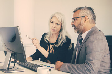 Wall Mural - Focused business colleagues watching content on computer, pointing at display and talking while sitting at desk with paper chart. Business communication concept