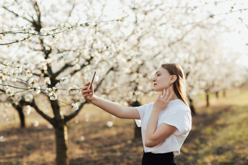 Portrait of a young beautiful girl enjoying spring in the park and making selfie. Blooming trees.