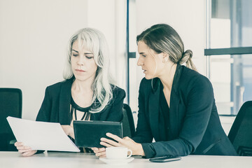 Wall Mural - Focused professionals analyzing reports together. Two businesswomen sitting together, reading documents, using tablet and talking. Teamwork concept