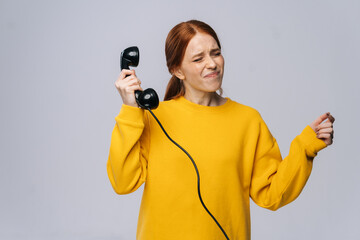 Unhappy young woman in stylish yellow sweater holding handset receiver of retro phone and turning head away from it, not wanting to hear what is being said to her.