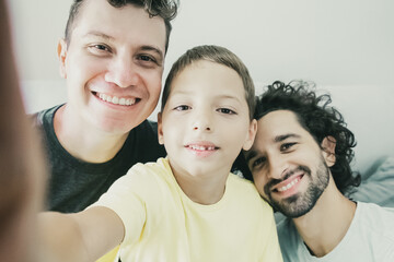 Selfie of cheerful homosexual dads and kid sitting on couch at home and smiling at frontal camera. Front view. Family and communication concept