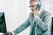© Mangostar - Serious grey haired CEO talking to cellphone and using computer at workplace in office. Medium shot. Communication and multitasking concept