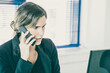 © Mangostar - Serious businesswoman in jacket speaking on mobile phone while using computer at workplace in office. Medium shot. Digital communication and multitasking concept
