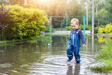 Wall Mural - Little cute playful caucasian blond toddler boy enjoy have fun playing jumping in dirty puddle wearing blue waterproof pants and rubber rainboots at home yard street outdoor. Happy childhood concept