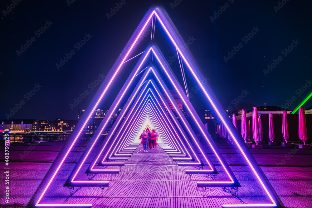 Purple coloured gate of light or purple light tunnel installation made of triangular neon and led lights at night as blurred people walk through the bright deep passage that resembles a human trachea - obrazy, fototapety, plakaty 