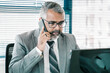 © Mangostar - Serious grey haired business professional in suit talking on cell phone while using computer at workplace in office. Medium shot. Digital communication and multitasking concept