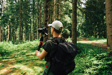 Wall Mural - Young male hiker in casual clothes stands in a mountain forest with a camera on a stabilizer and makes a video. Video operator captures content on camera during th