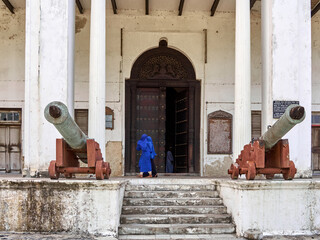 Two girls in blue burqa walking against old door and two powder canon's in front of white building. Zanzibar, Tanzania.