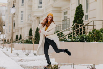 Wall Mural - Debonair girl in white coat jumping in winter day. Outdoor photo of dreamy woman enjoying cold weather.