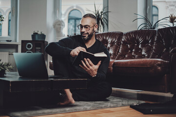 Portrait of a joy caucasian guy writing on notebook copying from laptop and sitting on floor in luxurios living room.