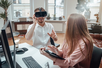 Bearded man in white shirt uses virtual headset and smiles while his female assistant with long brown hairs works on tablet in office room.