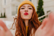 Ecstatic european girl in yellow hat posing in winter. Elegant ginger female model making selfie with kissing face expression.