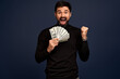 © Roman - Portrait of an excited young man in ttrendy black sweater holding bunch of money banknotes and celebrating isolated over Pacific Blue background