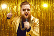 Funny singer in shiny golden jacket and gold chain holding microphone and singing songs at retro pop music concert. Happy bearded man performing at disco nightclub or enjoying time at karaoke party