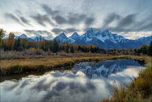 Autumn Evening Shot Of The Snow Covered Teton Mountains Reflected In Calm Water At Schwabachers Landing Of Grand Tetons National Park