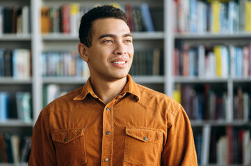 Portrait of a satisfied male freelancer or student. Confident successful hispanic business man looks away and smiling, standing in modern office or university library