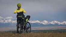 The Woman Travel On Mixed Terrain Cycle Touring With Bikepacking. The Traveler Journey With Bicycle Bags. Sport Tourism Bikepacking, Bike, Sportswear In Green Black Colors. Mountain Snow Capped.
