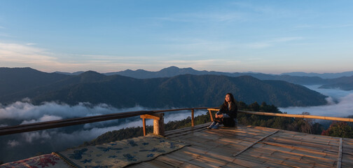 A young female traveler sitting on wooden balcony, looking at a beautiful mountain and sea of fog