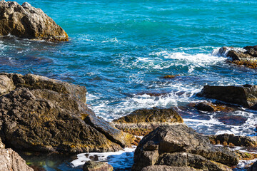 Wall Mural - Turquoise sea stone beach, breaking waves in bright sunlight. Colorful bright sea background. The concept of summer, vacation, travel. The purest clear sea water, large stones on the beach close-up