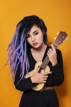 Beautiful Young Woman With Lilac Dreadlocks And Multi-colored Makeup Stands On A Yellow Studio Background And Playing An Ukulele