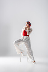 Wall Mural - full length of young woman in headscarf and stylish sunglasses sitting on stool and posing on white