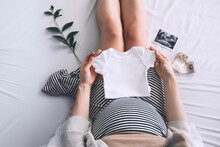 Pregnant Woman Holding White Baby Bodysuit Preparing To Child Birth During Pregnancy.