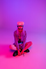 Poster - young woman in virtual reality headset sitting with crossed legs on purple and pink