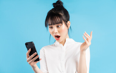 Wall Mural - Beautiful asian woman holding phone over blue background with surprised expression