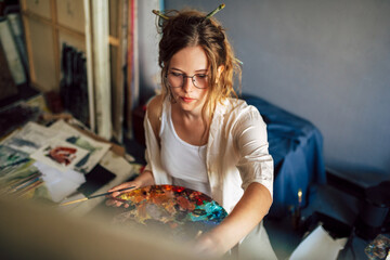 Wall Mural - Artist female sitting next to the easel with canvas painting something in her art studio. A professional candid young woman painter in transparent eyeglasses draws in the workshop.