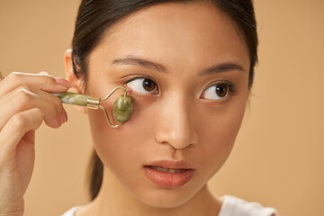 Wall Mural - Portrait of young woman looking aside while using jade roller for massaging her face, posing isolated over beige background