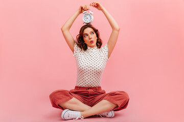 Curious girl with clock sitting on floor. Interested young woman posing on pink background.