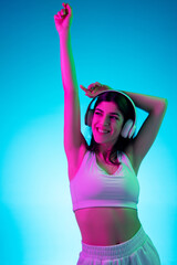 Wall Mural - Dancing. Brunette woman's portrait on blue studio background in mixed neon. Beautiful model with headphones listens to music. Concept of human emotions, facial expression, sales, ad, fashion.