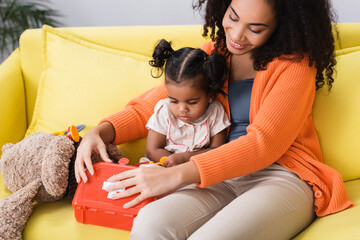 Wall Mural - happy african american mother sitting with toddler daughter and holding toy first aid kit in living room
