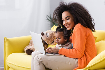 Wall Mural - smiling african american mother using laptop near toddler daughter in living room