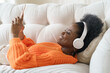 © DimaBerlin - African millennial woman in orange sweater wearing wireless headphones, listening music, holding and using smart phone, resting and lying on couch in living room. Girl spend lazy weekend at home.