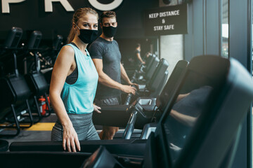 Young fit woman and man running on treadmill in modern fitness gym. They keeping distance and wearing protective face masks. Coronavirus world pandemic and sport theme.