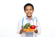 Young African American Child, Dressed As A Doctor, Recommends Vegetables And Fruits For Proper Nutrition And Benefits To Your Health