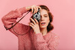 © Look! - Concentrated curly woman taking photos on pink background. Studio shot of charming girl with camera.