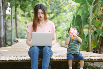 Portrait of young Asian mother and little son sit together on the floor in garden while mom working remotely on laptop and son playing with soap bubbles machine. Childcare and work concept.