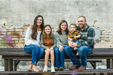 Blended Family Of Five With Two Girls And A Baby Boy Sitting On A Table By An Urban Old Brick Wall
