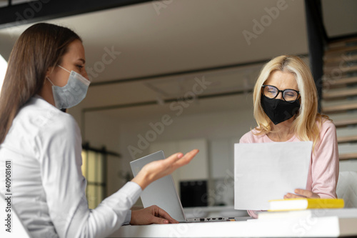 Confident female hr manager sitting at the office, wearing face mask, training young female worker intern, interviewing job seeker, mentoring a student. Social distance and safety at work concept