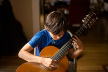 A Boy Hunches Over A Classical Guitar Playing A Chord In Window Light