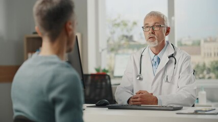 Wall Mural - Middle Aged Family Doctor is Talking with Young Male Patient During Consultation in a Health Clinic. Senior Physician in Lab Coat Sitting Behind a Computer Desk in Hospital Office.