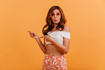 Canvas Print - Photo of girl on orange background with place for text. Lady in pink glasses is looking at camera in surprise and pointing with her fingers to left