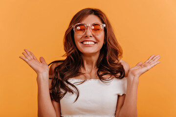 Close-up portrait of insanely happy girl covering her eyes with joy. Lady in white t-shirt and orange glasses laughs